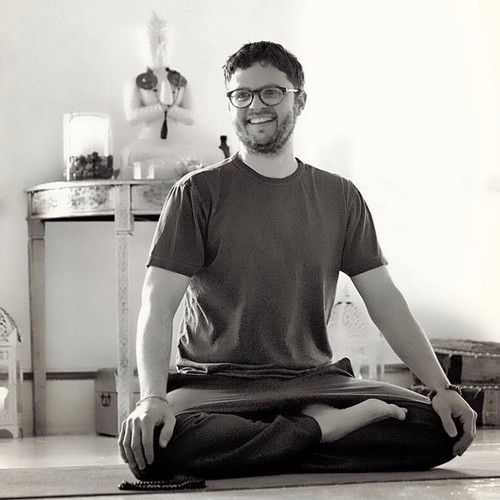 Black and white photo of bearded, young white man wearing glasses seated cross-legged on the floor. 