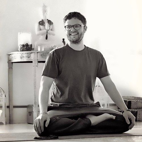 Black and white photo of bearded, young white man wearing glasses seated cross-legged on the floor.